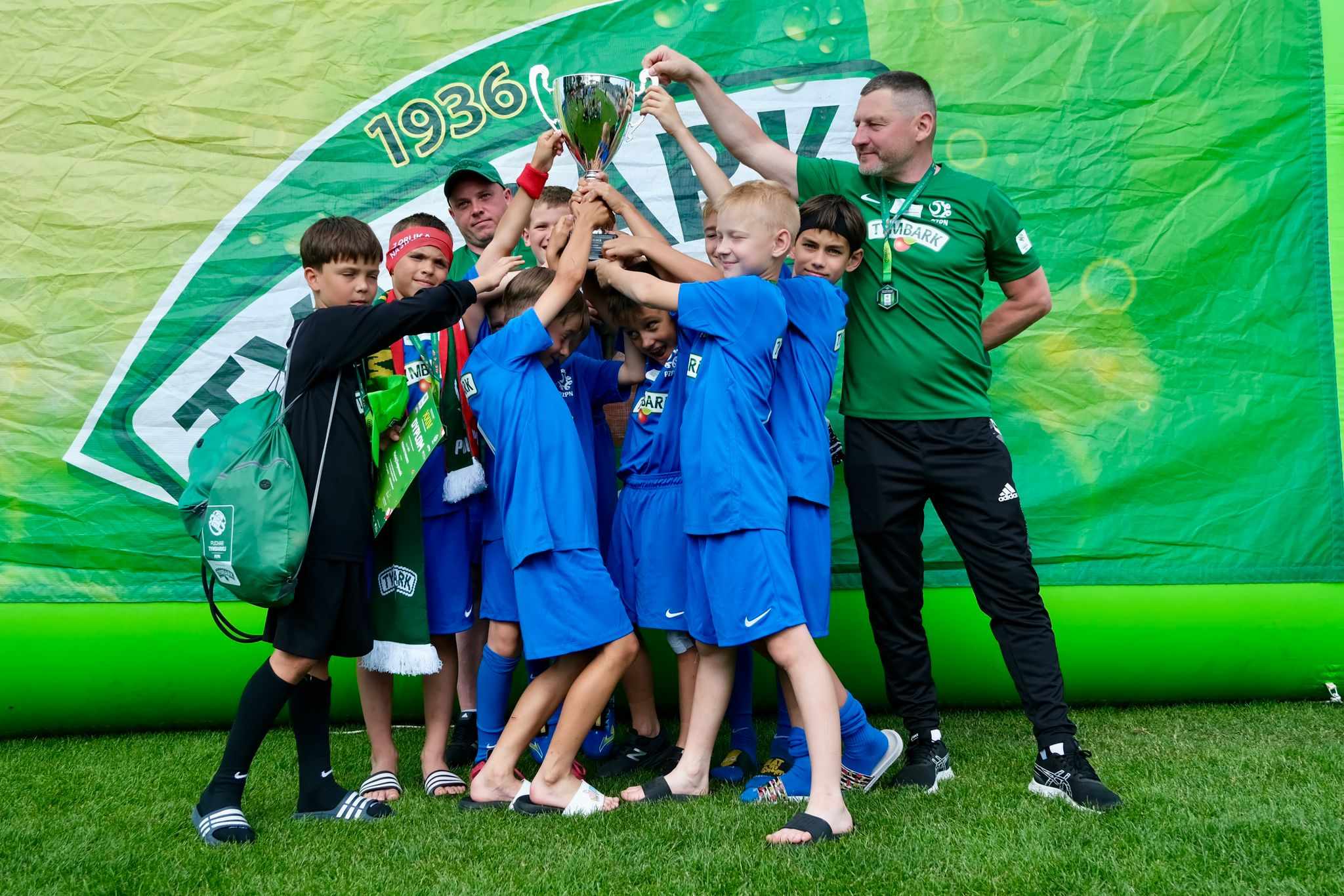 Grupa uczniów ubrana w niebieskie stroje sportowe trzyma puchar razem z trenerami. W tle zielony billboard..jpg
