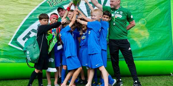 Grupa uczniów ubrana w niebieskie stroje sportowe trzyma puchar razem z trenerami. W tle zielony billboard..jpg
