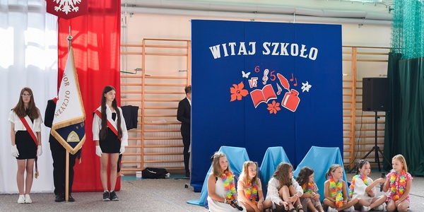 Troje uczniów stoi ze sztandarem na tle flagi polskiej, obok siedzi grupa dzieci z kwietnymi łańcuchami.
