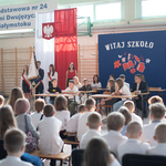 Widownia zgormadzona na sali patrzy na grupę uczniów siedzących w ławkach. W tle napis Witaj szkoło.