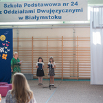 Dwie dziewczynki stoją przed mikrofonem, w tle flaga biało-czerwona .
