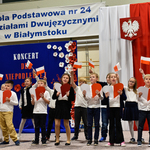 Grupa dzieci ubrana na galowo macha w górze flagami biało-czerwonymi.