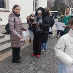 Grupa uczniów ubrana w kurtki stoi przed budynkiem z kartkami w rękach_..jpg